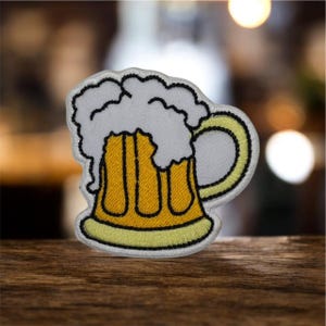 May include: Embroidered patch featuring a cartoon beer mug with a frothy white head and a yellow body. The mug has a curved handle and is outlined in black. The patch is designed to be ironed on or sewn onto fabric.