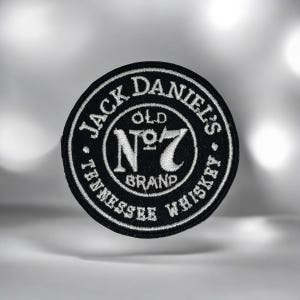 May include: A circular black embroidered patch featuring white stitched text that reads Jack Daniel's Old No. 7 Brand Tennessee Whiskey. The design includes a central emblem with the number 7 and decorative borders, creating a high-contrast look.
