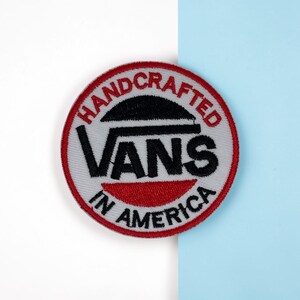 Pode incluir: Um emblema bordado circular com fundo cinza claro e borda vermelha. O texto HANDCRAFTED IN AMERICA está bordado em vermelho ao redor do perímetro, enquanto o logotipo preto da VANS está centralizado com uma barra horizontal preta acima.