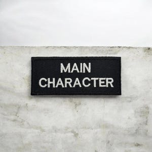 Puede incluir: Un parche rectangular negro con las palabras "MAIN CHARACTER" bordadas en blanco. El parche se muestra sobre un fondo texturizado de color claro.