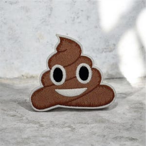 May include: Embroidered brown poop emoji patch with a smiling face, black eyes, and a white mouth. The patch has a white border, suitable for clothing or accessories.