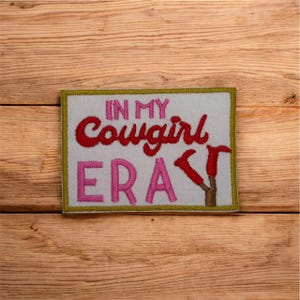 Pode incluir: Patch bordado com o texto "IN MY Cowgirl ERA" em letras vermelhas e rosa. O patch tem um fundo cinza claro com uma borda dourada e um design de bota marrom. O patch é retangular.