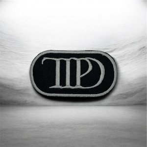 May include: Black and gray embroidered patch with the letters "TP" in a stylized font. The patch has a black background and a gray border and lettering. The patch is oval-shaped.