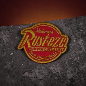 Puede incluir: Un parche bordado circular con fondo rojo y costuras doradas. El texto dice Medicated Rust-eze Bumper Ointment Rear End Formula en una fuente estilizada. El parche tiene un borde dorado y una forma distintiva con un lado puntiagudo.