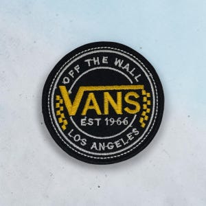May include: A circular black embroidered patch featuring the yellow Vans logo in the center. The text reads OFF THE WALL, VANS, EST 1966, and LOS ANGELES. The design includes a yellow and black checkered pattern on the sides of the logo.