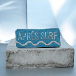 May include: A turquoise rectangular patch with white embroidered text reading "APRÈS SURF", above a white wave design. The patch is on a concrete surface against a light blue backdrop.