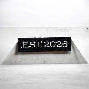 Puede incluir: Un parche rectangular negro con texto bordado en blanco ".EST. 2026". El parche está centrado y parece estar hecho de un material similar al fieltro. El fondo es una superficie blanca texturizada.