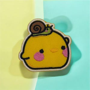 May include: Embroidered patch featuring a yellow cartoon character with pink cheeks and a small snail on its head. The patch has a black outline and a tan border. The background is split between yellow and teal.