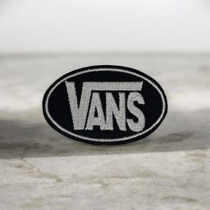 Könnte beinhalten: Ein ovaler, bestickter Aufnäher mit schwarzem Hintergrund, weißem Rand und dem Wort VANS, das in weißen Großbuchstaben in der Mitte aufgestickt ist. Das Design zeigt das charakteristische V, das über die Buchstaben A, N und S reicht.
