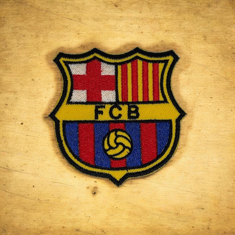 Op de afbeelding: Een geborduurde patch met het embleem van FC Barcelona, voorzien van een rood kruis op een witte achtergrond, gele en rode verticale strepen, een gele centrale band met de zwarte tekst FCB en een gele voetbal op blauwe en rode strepen.