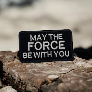 May include: A black rectangular patch with white embroidered text that reads "MAY THE FORCE BE WITH YOU." The patch is set against a blurred, neutral background, suggesting an outdoor setting. The patch is likely an accessory.