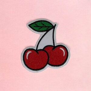 May include: An embroidered patch featuring two round red cherries with white highlights and a single green leaf. The design is outlined in black thread with a light grey border, creating a distinct shape against the solid background.