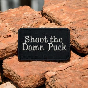 Op de afbeelding: Een zwart rechthoekig embleem met witte geborduurde tekst die luidt "Shoot the Damn Puck". De patch staat tegen een achtergrond van ruwe, roodbruine bakstenen.