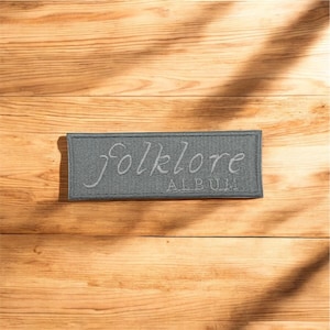 May include: A rectangular gray patch with the word "Folklore" in cursive script above the word "Album" in a sans-serif font. The patch is on a wooden surface with a natural wood grain.