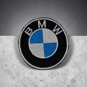 Pode incluir: Um emblema bordado circular com o logótipo da BMW. O design inclui um anel exterior preto com as letras brancas BMW e um círculo central dividido em quatro quadrantes com cores alternadas azul e branco prateado.
