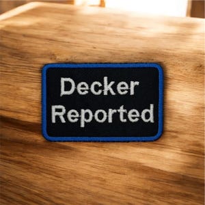 Pode incluir: Um patch retangular com as palavras "Decker Reported" bordadas em branco sobre um fundo preto. O patch tem uma borda azul e está sobre uma superfície de madeira.