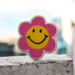 Puede incluir: Parche bordado de flores con una cara sonriente. El parche presenta una flor rosa con un centro amarillo y una cara sonriente negra. La flor tiene un borde blanco.