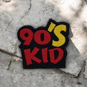 Puede incluir: Un parche bordado negro con las palabras "90'S KID" en letras rojas y amarillas. El parche tiene un borde negro y está diseñado para ser planchado o cosido en ropa o accesorios.