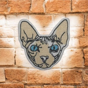 May include: Embroidered patch depicting a Sphynx cat's face with bright blue eyes. The patch has a tan and grey colour scheme with detailed stitching and a grey border. This patch is suitable for adding a unique touch to clothing or accessories.