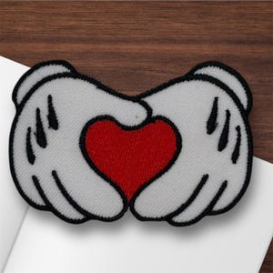 May include: An embroidered patch featuring white cartoon gloves forming a heart shape with a red heart in the center. The gloves have black outlines and details. The patch is on a white surface with a wooden background.