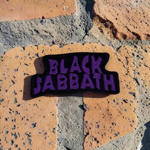 May include: A rectangular embroidered patch with a black background and the words BLACK SABBATH stitched in a bold, purple, stylized font. The patch features a thick black border and a distinct, uneven shape that follows the lettering contour.
