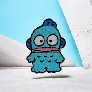 Puede incluir: Un parche bordado que muestra un personaje de pez de dibujos animados con piel azul brillante, ojos grandes blancos con pupilas negras y una boca ancha de color rosa. El personaje tiene aletas pequeñas a los lados y una cola festoneada.