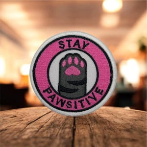Può includere: Una toppa rotonda ricamata con un bordo rosa e le parole "STAY PAWSITIVE". La toppa presenta una zampa di gatto grigia con cuscinetti rosa all'interno di un cerchio bianco.