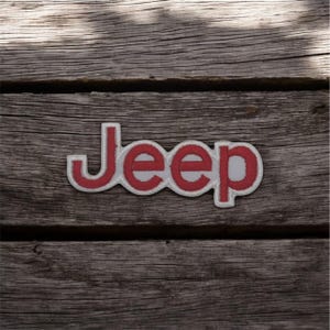 Peut inclure: Écusson brodé avec le mot "JEEP" en rouge et un contour blanc. L'écusson est posé sur une surface en bois grisé et vieilli. Idéal pour les vêtements ou accessoires.