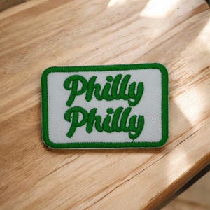 May include: A rectangular patch with a white background and a green border. The patch features the words "Philly Philly" in green, cursive lettering. The patch is likely an embroidered item.