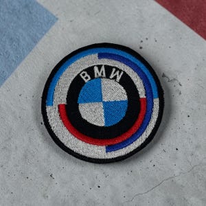 Puede incluir: Un parche bordado circular que presenta el logotipo de BMW en el centro con cuadrantes blancos y azules. El emblema está rodeado por un anillo negro con el texto blanco BMW en la parte superior, rodeado por un borde segmentado plateado, azul y rojo.