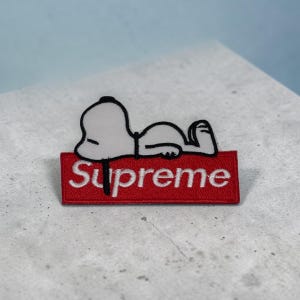 May include: An embroidered patch featuring a white cartoon dog lying down on top of a rectangular red box. The red box contains the white text Supreme in a bold, sans-serif font. The patch has a black stitched outline around the dog figure.