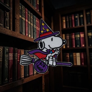 Puede incluir: Un parche bordado que muestra a un perro de dibujos animados blanco con un sombrero de mago morado con estrellas amarillas y una luna creciente, una capa morada con forro rojo, montado en una escoba morada con una calabaza morada.