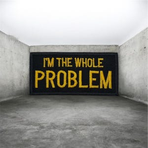 Op de afbeelding: Zwart rechthoekig embleem met gele geborduurde tekst "I'M THE WHOLE PROBLEM". Het embleem staat tegen een grijze betonnen achtergrond.