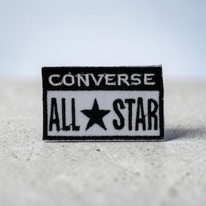 May include: A rectangular embroidered patch featuring a black border and a black top section with the white text CONVERSE. The bottom section is white with black text reading ALL STAR and a black star symbol centered between the two words.