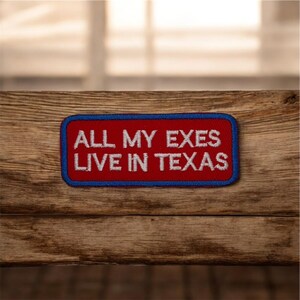 Peut inclure: Un écusson rectangulaire avec un fond rouge et une bordure bleue. Le texte "ALL MY EXES LIVE IN TEXAS" est brodé en blanc. L'écusson est posé sur une surface en bois.