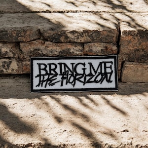 Könnte beinhalten: Ein rechteckiger, bestickter Aufnäher mit weißem Hintergrund und einem dicken schwarzen Rand. Der Text "BRING ME THE HORIZON" ist in schwarzem Garn mittig in einer stilisierten, zackigen und abgenutzten Schriftart aufgestickt.