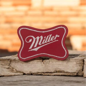 May include: A red and white embroidered patch with the word "Miller" in a cursive font. The patch has a unique shape with a white border and is set against a rustic wooden surface.