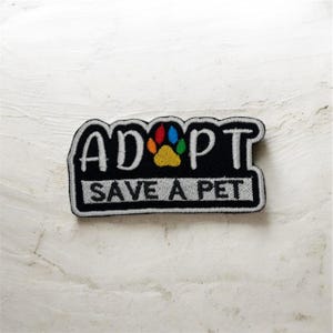 Puede incluir: Parche bordado negro con las palabras "ADOPT" y "SAVE A PET" en letras blancas. Un diseño de huella de pata colorida está en el centro. El parche es rectangular con un borde blanco.