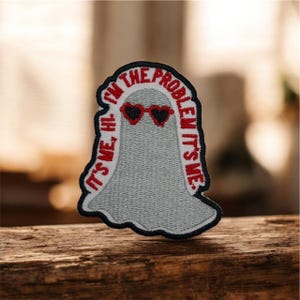 Pode incluir: Patch bordado com um fantasma cinzento a usar óculos de sol em forma de coração. O patch tem o texto "I'M THE PROBLEM IT'S ME" em vermelho, com "IT'S ME, HI" à esquerda. O patch tem uma borda preta.