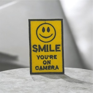 Puede incluir: Un parche rectangular amarillo con un borde negro y las palabras "SMILE YOU'RE ON CAMERA" en negro. Encima del texto hay una carita sonriente negra. El parche es probablemente de tela y se muestra sobre una superficie gris claro.