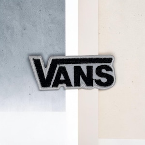 Peut inclure: Un écusson brodé rectangulaire présentant le texte noir VANS sur un fond gris clair. L'écusson a une forme distincte qui suit le contour des lettres, avec une bordure grise épaisse entourant le logo brodé en noir.