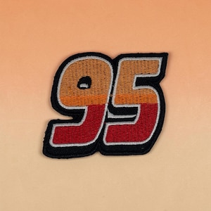 May include: An embroidered patch featuring the number 95 in a bold, block-style font. The design displays a horizontal gradient transitioning from orange at the top to red at the bottom, outlined with a thick black border and a thin silver edge.