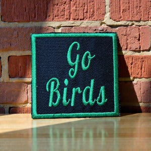 Puede incluir: Un parche cuadrado con las palabras "Go Birds" bordadas en verde sobre un fondo negro. El parche tiene un borde verde y está colocado contra una pared de ladrillos y una superficie de madera.