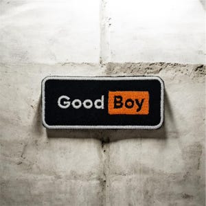 May include: A rectangular black patch with the words "Good Boy" in white and orange. The patch has a black border and is set against a textured, light-colored background.