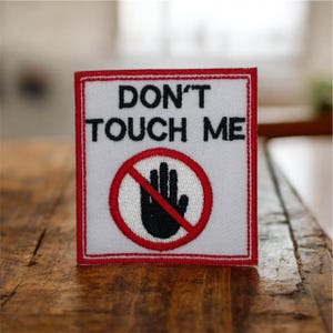 May include: Square embroidered patch with a white background, red border, and the words "DON'T TOUCH ME" in black. A black hand inside a red circle with a red slash through it is also featured.