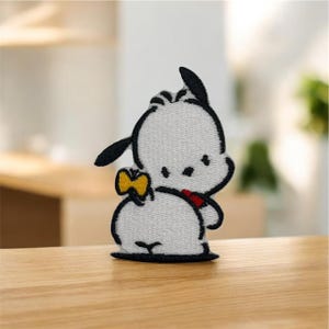 以下が含まれることがあります： 白い毛並み、黒い耳、黒い輪郭を持つ漫画の犬のキャラクターが刺繍されたパッチ。犬は黄色の蝶ネクタイと赤い襟をしています。パッチは木製の表面にあります。