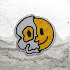 Può includere: Toppa ricamata con un design diviso: un teschio grigio a sinistra e una faccina sorridente gialla a destra. La toppa ha un bordo nero ed è posta su una superficie chiara e strutturata.