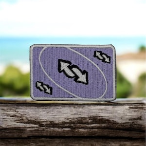 Puede incluir: Un parche rectangular con fondo morado claro y borde blanco. El parche presenta un óvalo blanco con dos flechas negras que apuntan en direcciones opuestas y tres flechas negras más pequeñas. El parche es probablemente para ropa o accesorios.