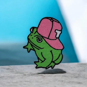 Puede incluir: Parche bordado con una rana verde que lleva una gorra de béisbol rosa con una "T" blanca. La rana tiene una expresión severa y está delineada en negro. Para ropa o accesorios.