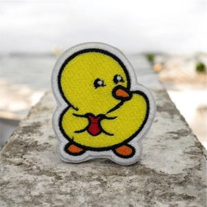 Puede incluir: Parche bordado con un patito de dibujos animados. El patito es amarillo con contorno blanco, ojos negros, pico y patas naranjas, y un lazo rojo. Para ropa o accesorios.